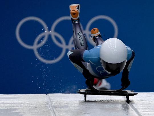 Katie Tannenbaum hopping onto a skeleton sled with the Olympics rings behind her.